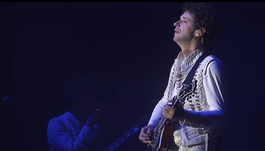 Gustavo Cerati en vivo con traje blanco durante la gira Fuerza Natural, tocando la guitarra bajo luces tenues.