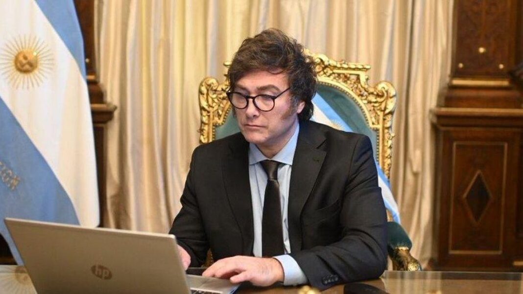 El presidente argentino Javier Milei en la Casa Rosada, vestido con traje y corbata, trabajando frente a una notebook HP, con la bandera argentina y cortinas doradas de fondo.
