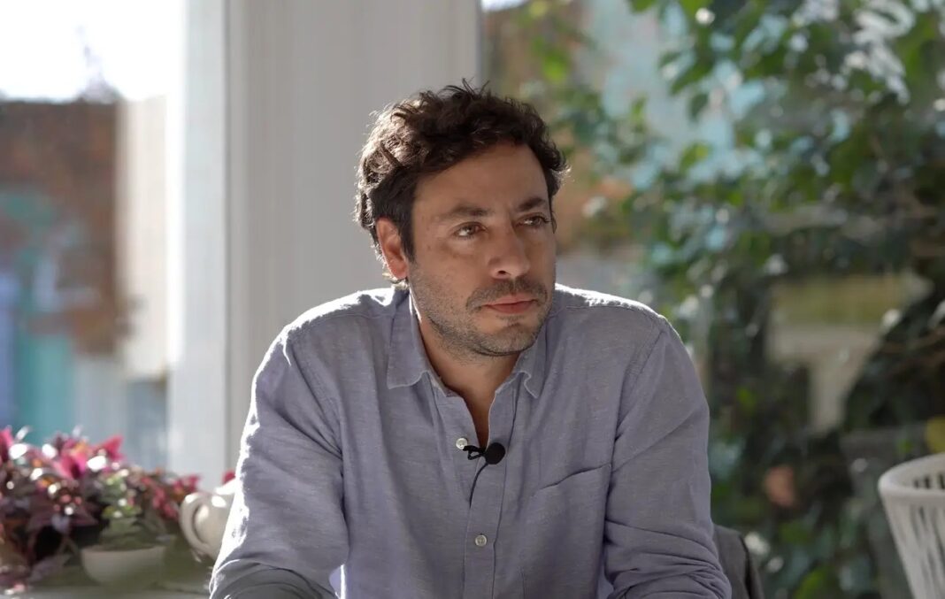 El intendente de Pinamar, Juan Ibarguren, durante una entrevista, con gesto serio y micrófono de solapa, en un ambiente interior con plantas de fondo.